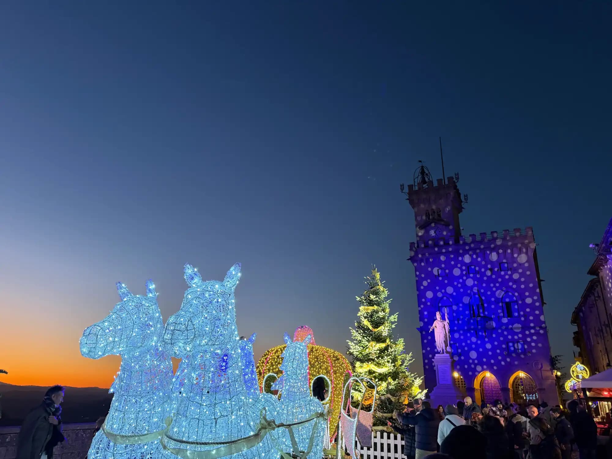 Nighttime town square with illuminated horse sculptures, a Christmas tree, and a historic tower lit in blue