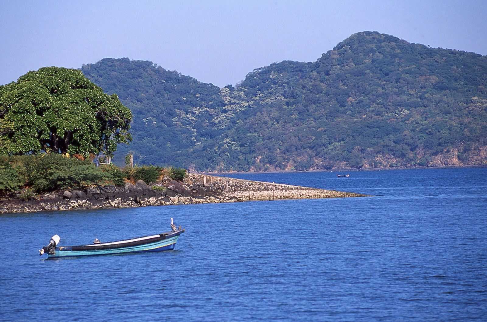 Coastal gulf shoreline with a small motorboat, rocky breakwater, lush hills, and calm blue water