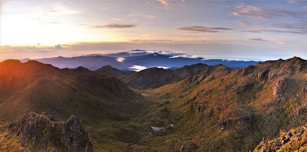 Mountain landscape with rolling ridges, distant peaks above a cloud layer, and warm sunset light.