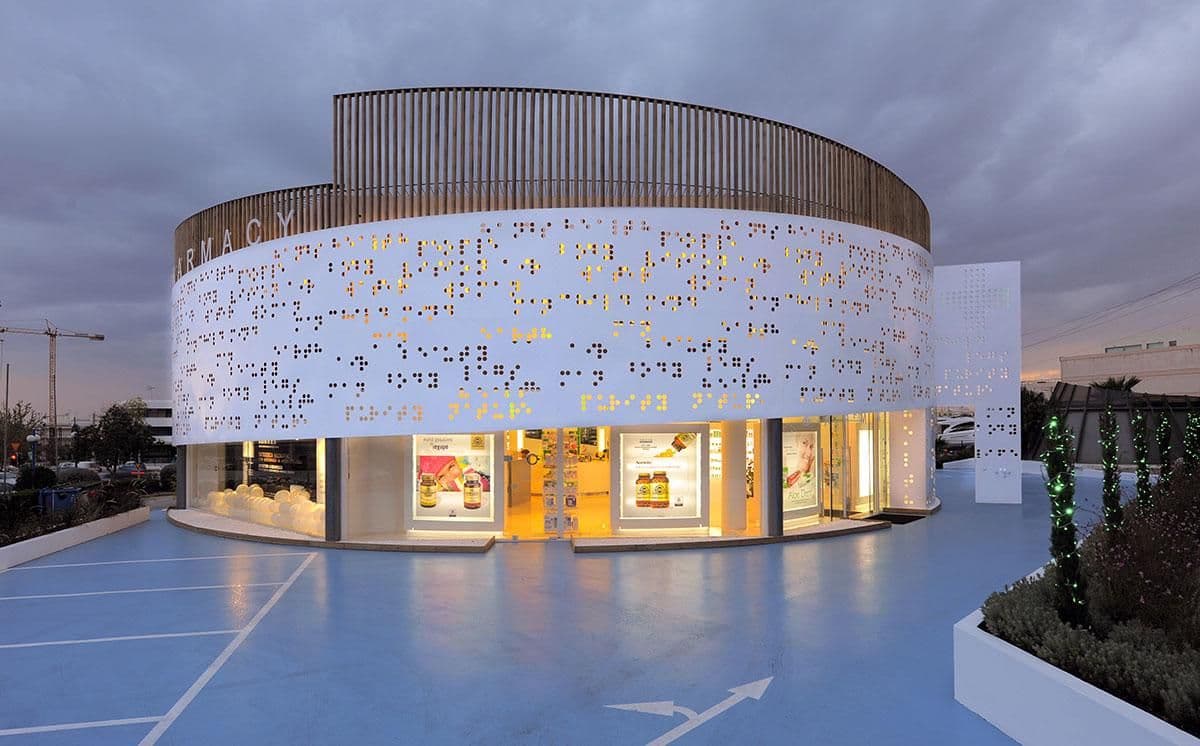 Exterior of a modern circular pharmacy building with perforated facade, illuminated storefront, and blue paved plaza at dusk.