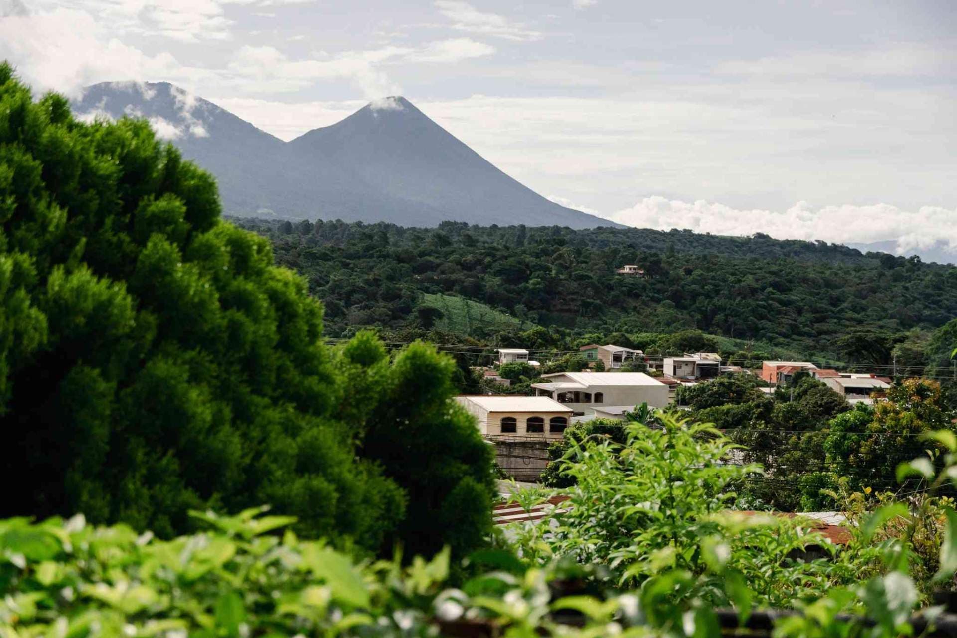 Lush green hills and a rural village with a volcano in the background under a cloudy sky.