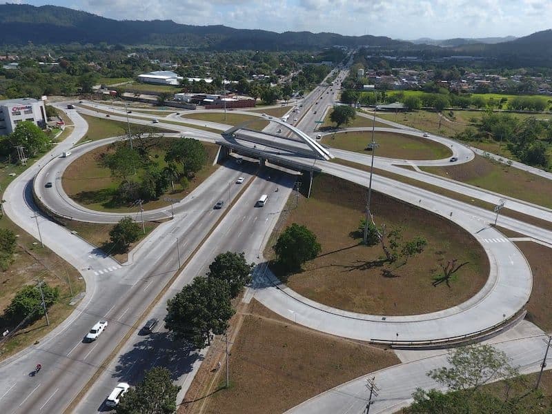 Aerial view of a multi-lane highway interchange with a pedestrian bridge, leading into a city with hills in the background.