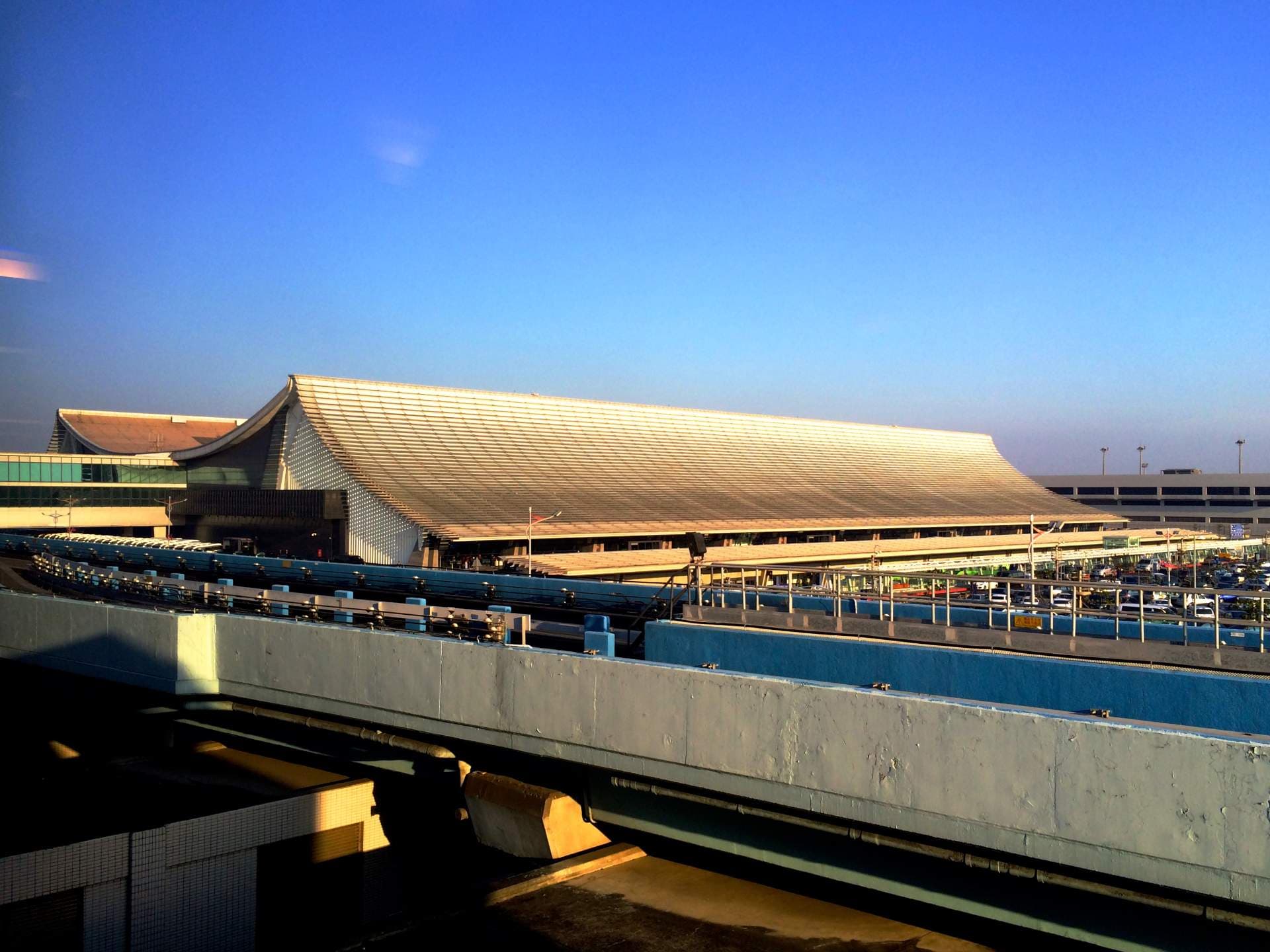 Exterior view of a large modern high-speed rail station with a distinctive sloped roof, elevated tracks, and clear blue sky.