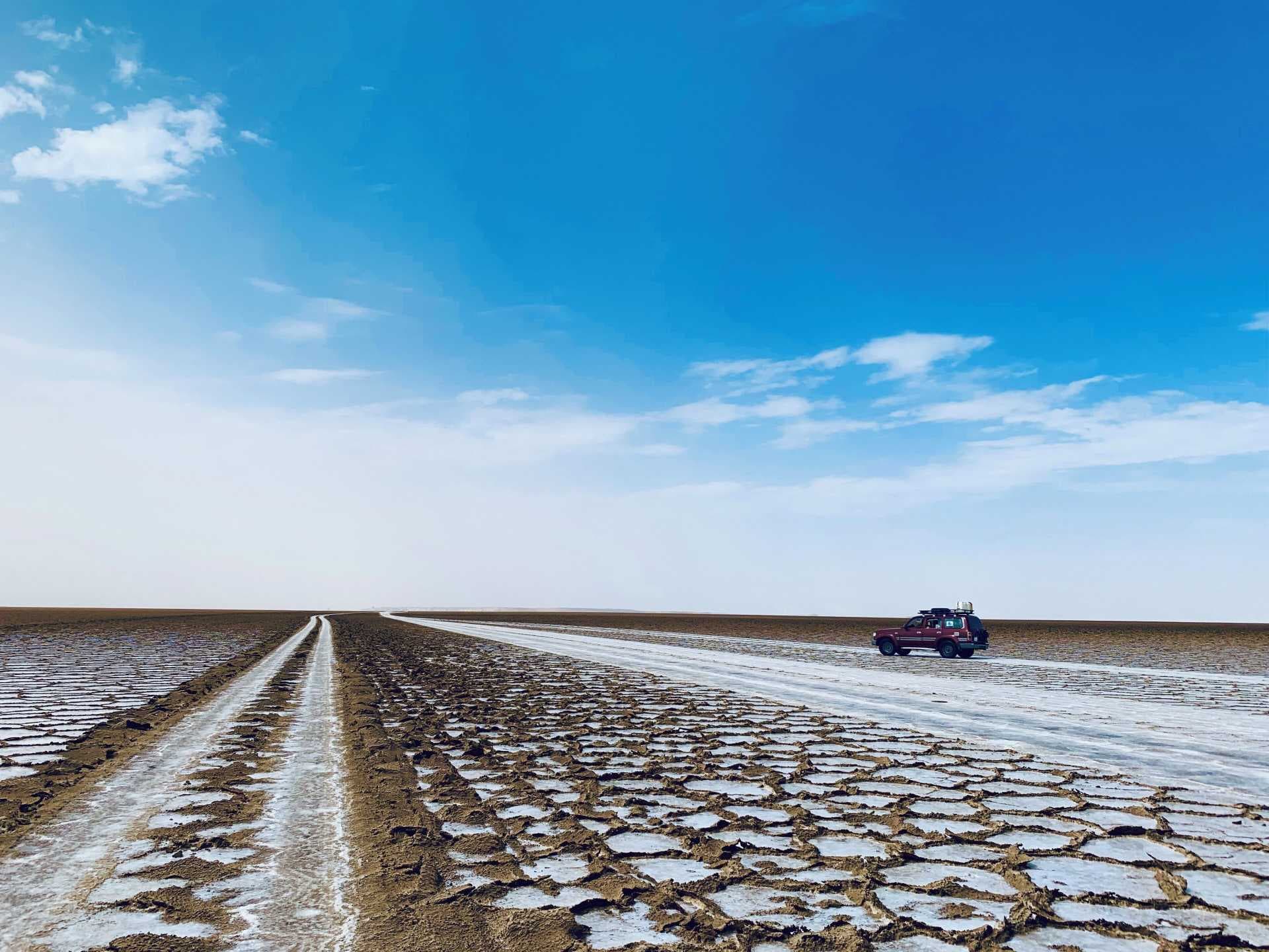A lone red SUV sits on a cracked, salt-encrusted road stretching across the arid Afar landscape under a bright blue sky.