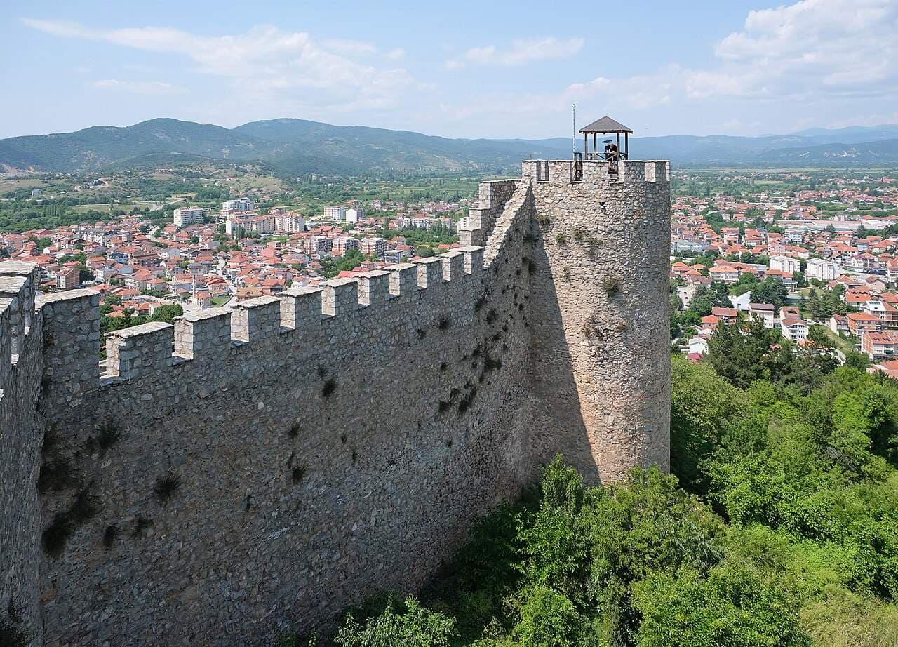 Stone fortress wall and tower overlooking a red-roofed town with hills and a distant lake under a partly cloudy sky.