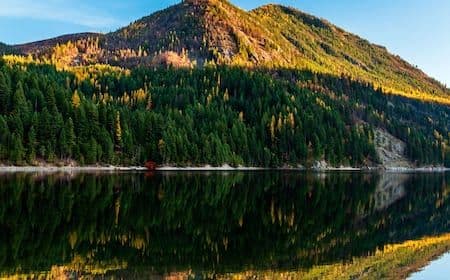 A forested mountain peak with autumn foliage reflected in a calm lake near the Colville area, captured in warm golden light.