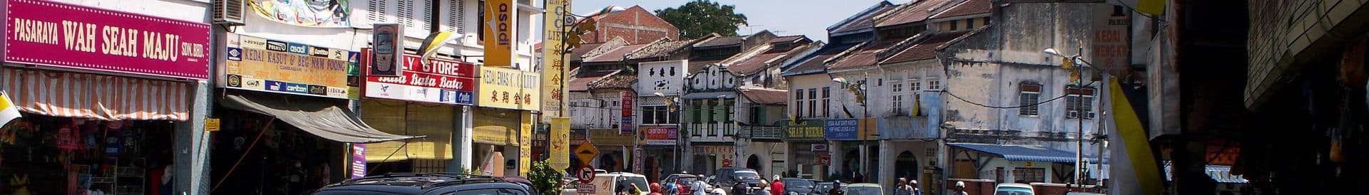 Wide streetscape of a Malaysian old town with shophouses, colorful shop signs, parked vehicles, and a few pedestrians