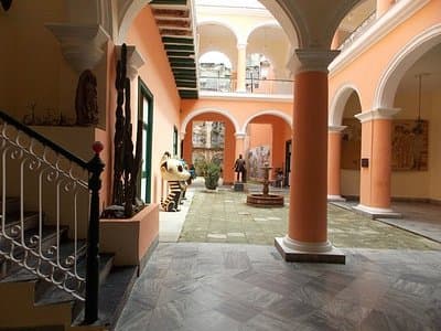 Interior courtyard of a peach-colored building featuring arched walkways, columns, and a staircase, with a few people visible in the background.