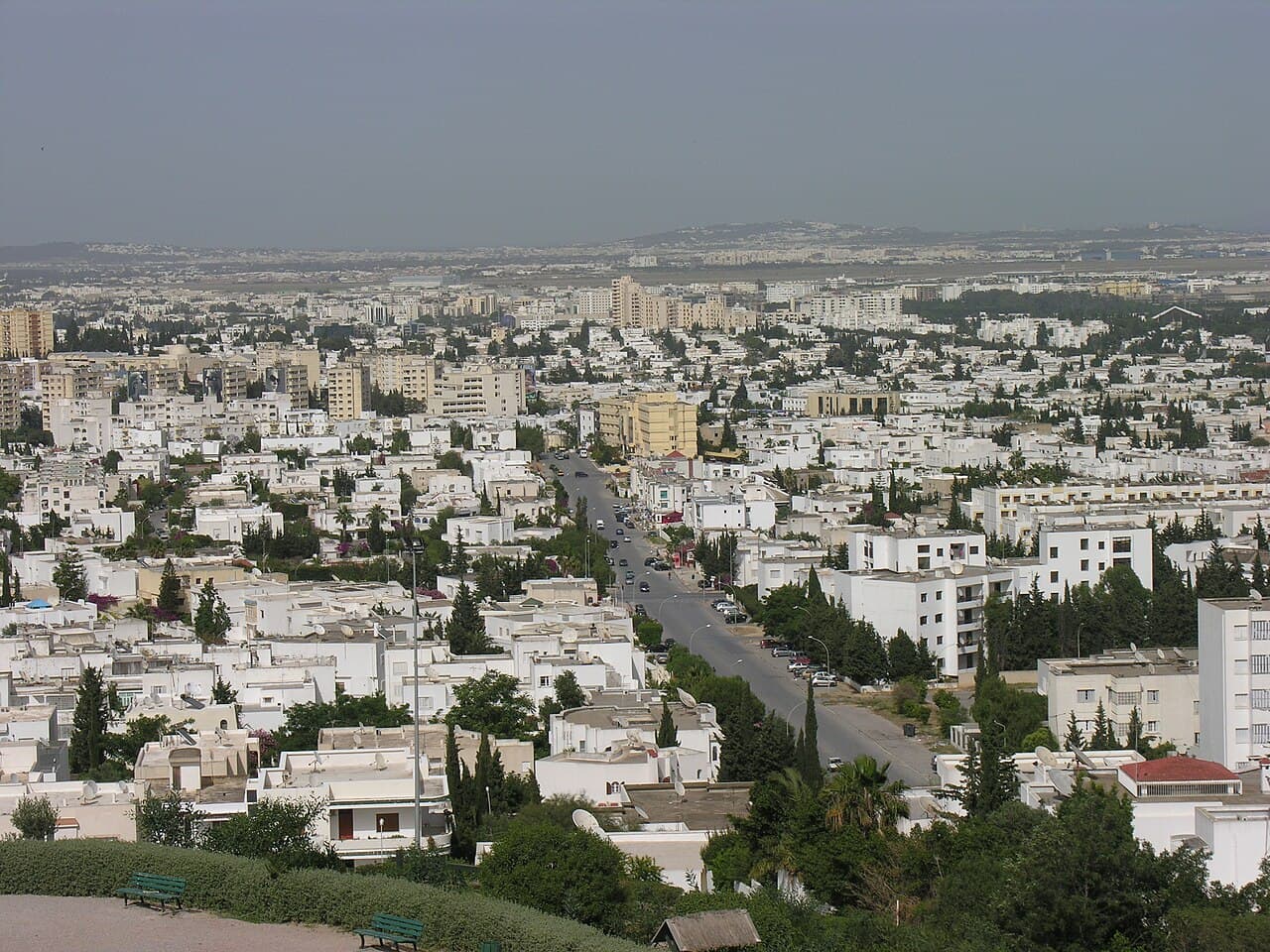 Aerial view of Ben Arous showing dense white residential apartment buildings, scattered greenery, and a main road stretching toward the horizon.