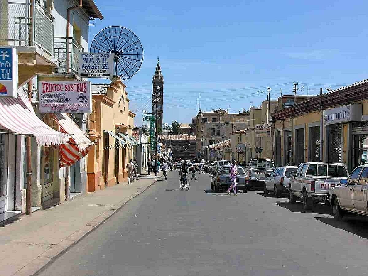 Street scene in Adi Keyh, Eritrea, with local buildings, parked vehicles, a cyclist, and a clock tower in the distance.