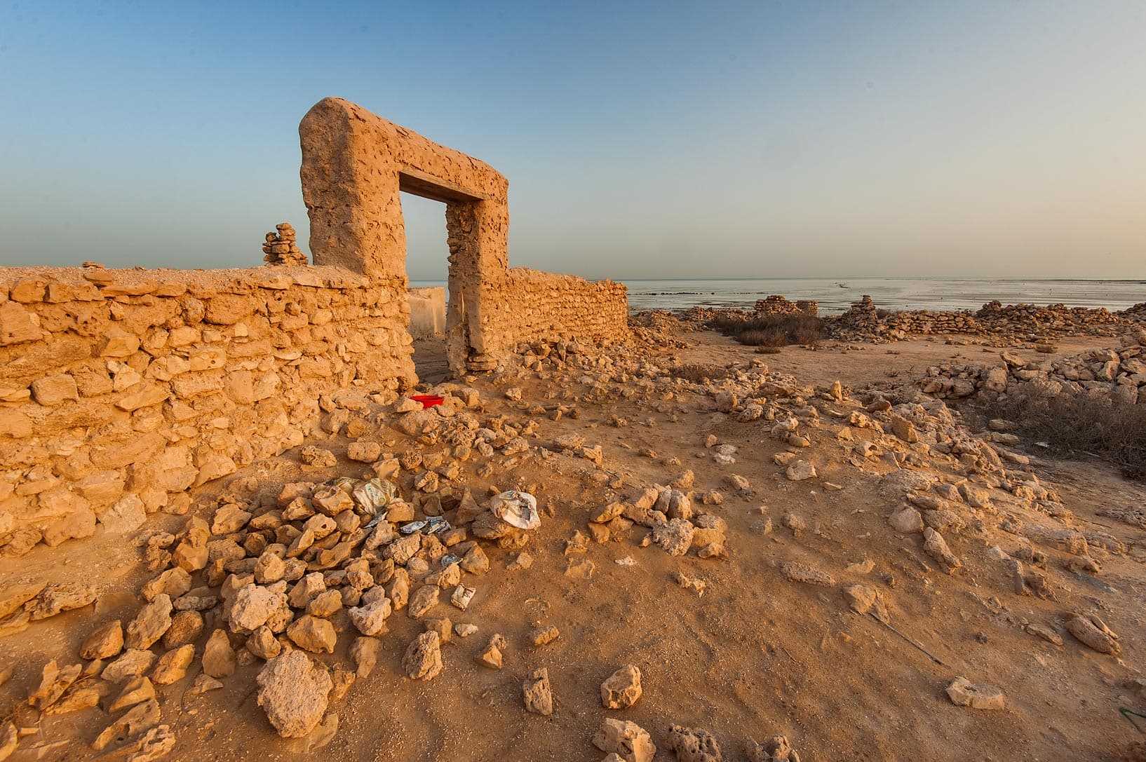Sunlit ruins of a stone structure with a doorway frame stand in a desert landscape near the sea horizon.