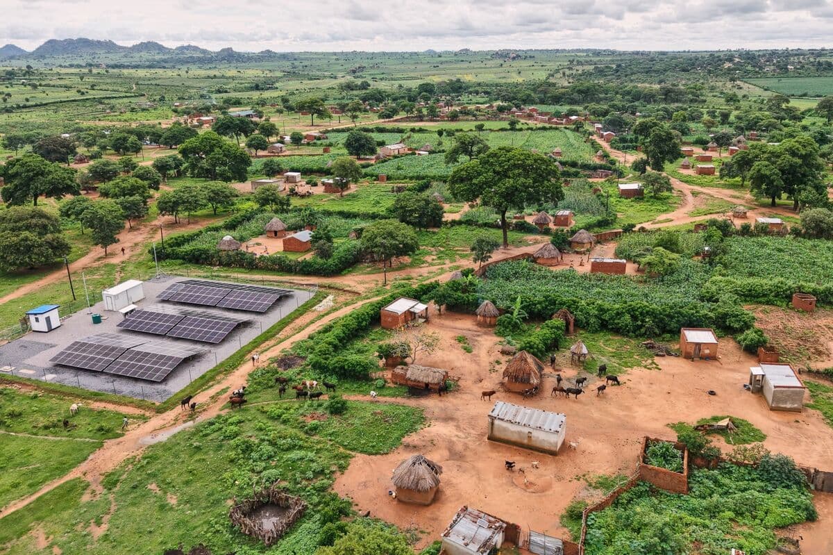 Aerial view of a rural Zambian village with thatched huts, solar panels, livestock, and green farmland under a cloudy sky.