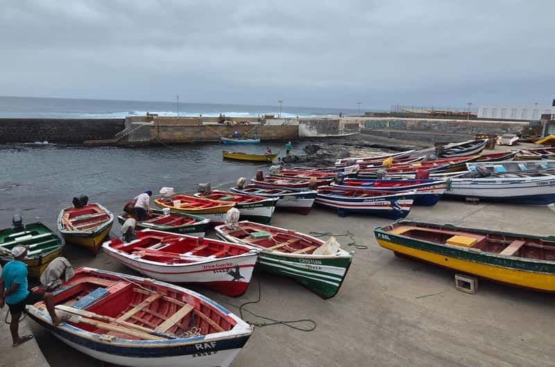 Colorful fishing boats rest on a concrete harbor shore under overcast skies, with a stone breakwater and ocean waves visible in the background.