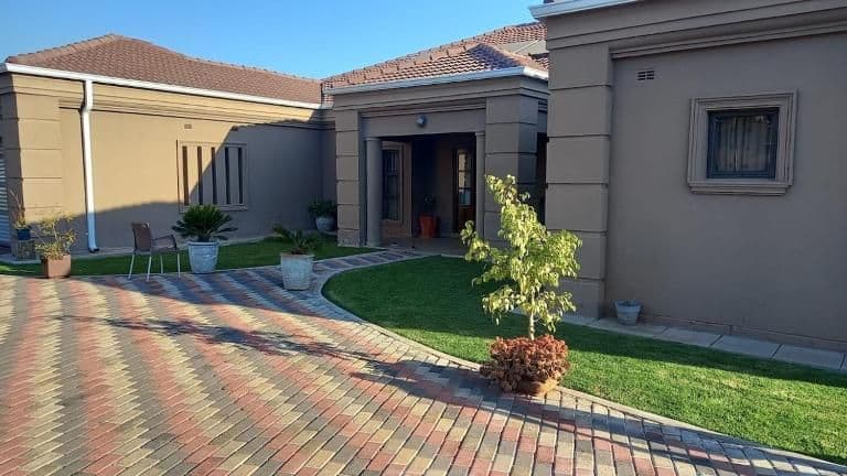 Exterior of a single-story guesthouse in Marondera, Zimbabwe with a paved courtyard, lawn, potted plants, and tiled roof under a clear sky