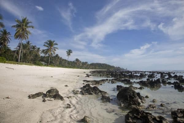 A wide-angle view of a tropical beach featuring white sand, scattered dark rocks, and palm trees under a blue sky with wispy clouds in Anabar, Nauru.