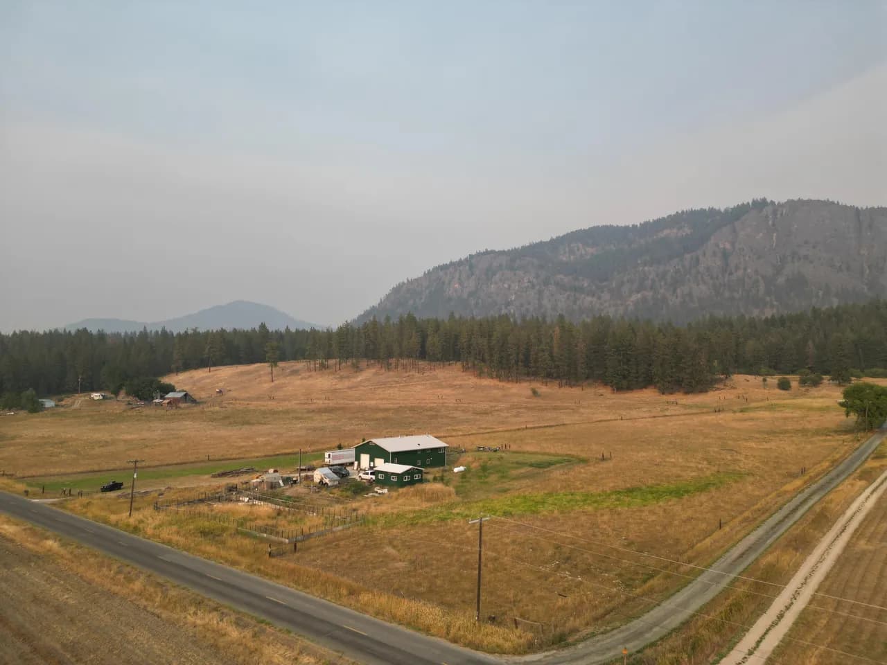 Aerial view of a rural two-lane road crossing dry fields near Chewelah, Washington, with a green barn, forested hills, and distant mountains.