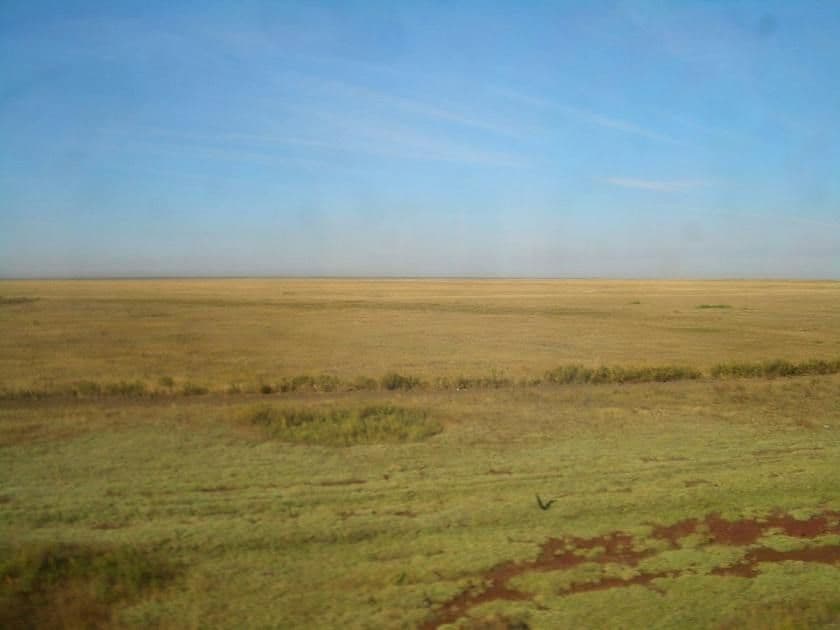 Wide flat Kazakh steppe landscape under a clear blue sky with sparse low vegetation and a distant horizon.