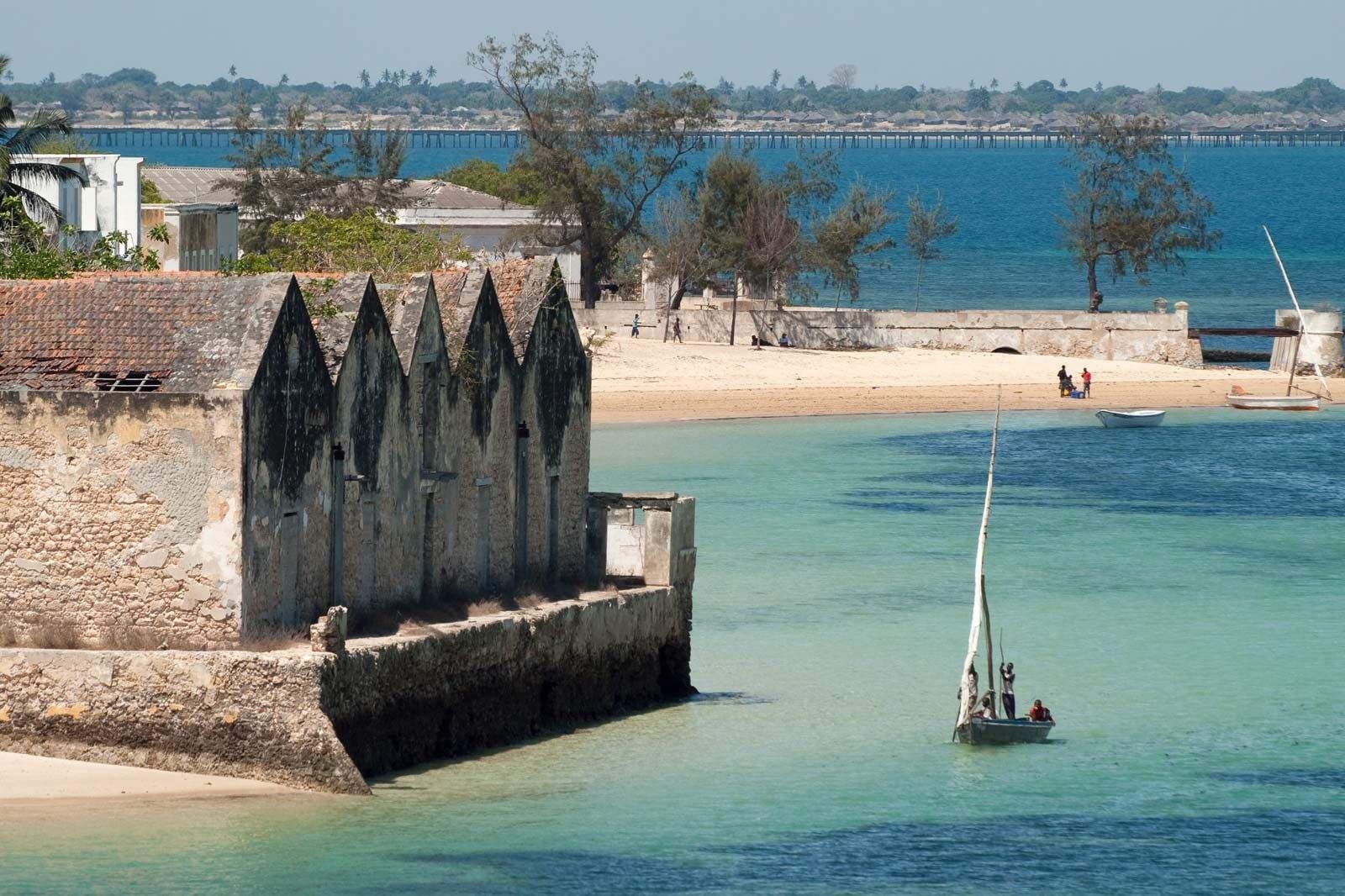 Coastal view featuring historic stone fortifications, turquoise water, and a traditional dhow sailboat near a sandy beach.
