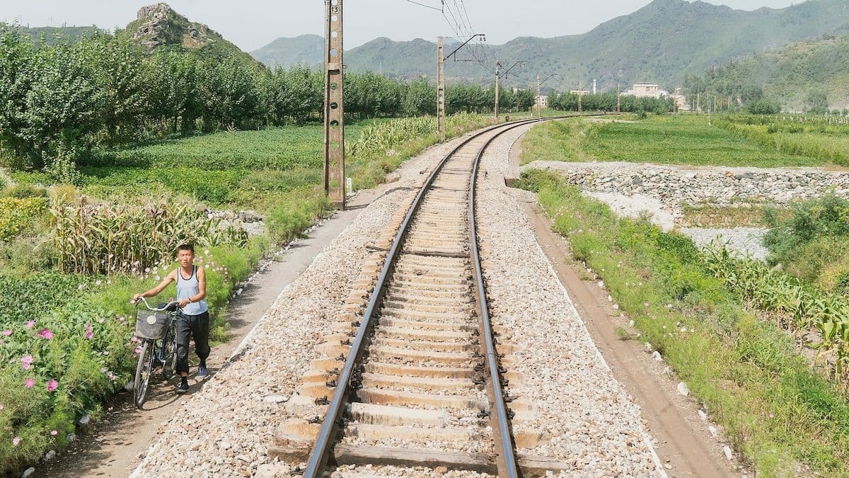 Rural railway track curving through green fields and hills, with a boy on a bicycle beside the line.