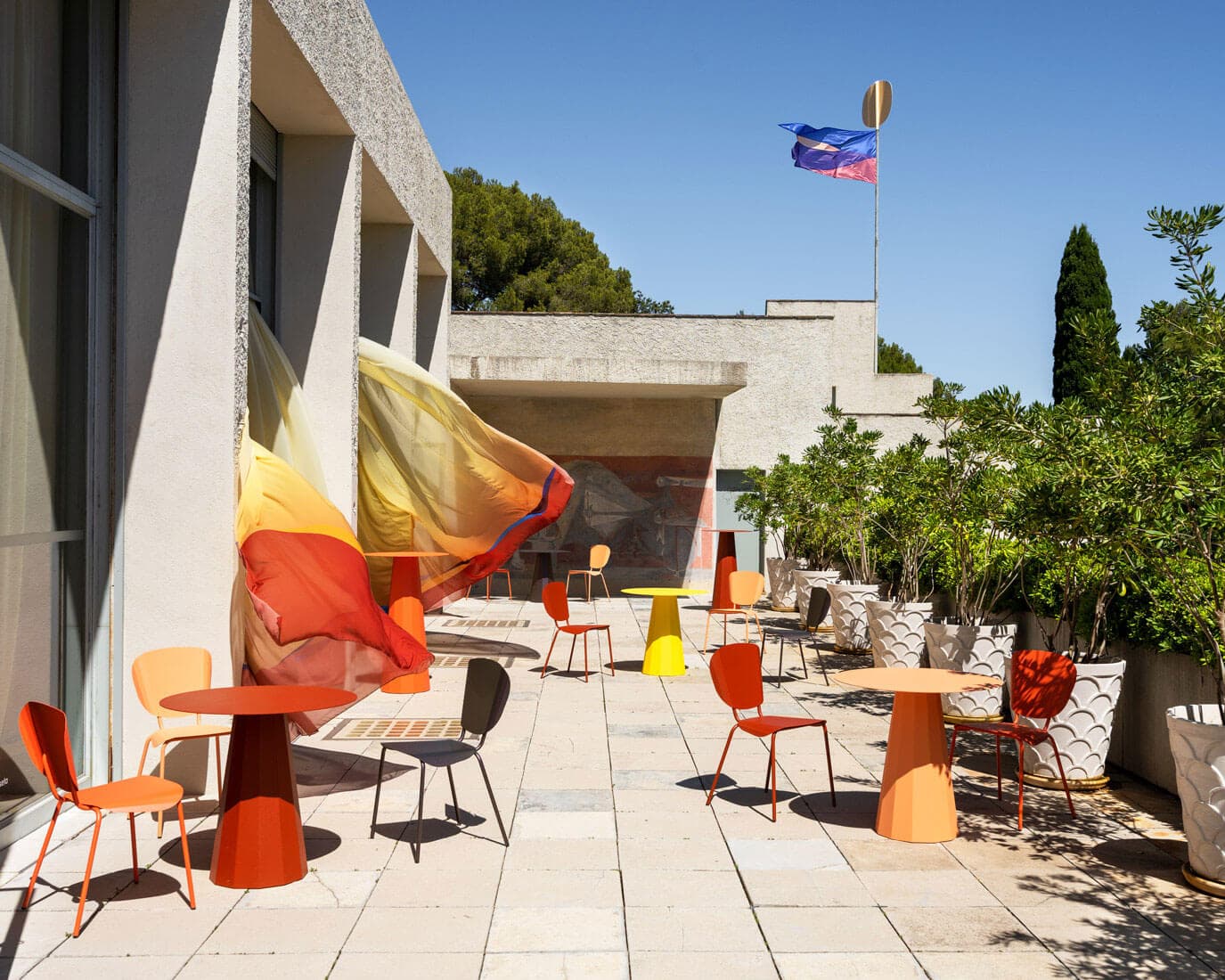 Colorful chairs and tables arranged on a sunny terrace with modern architecture, potted plants, and a flag against a blue sky.