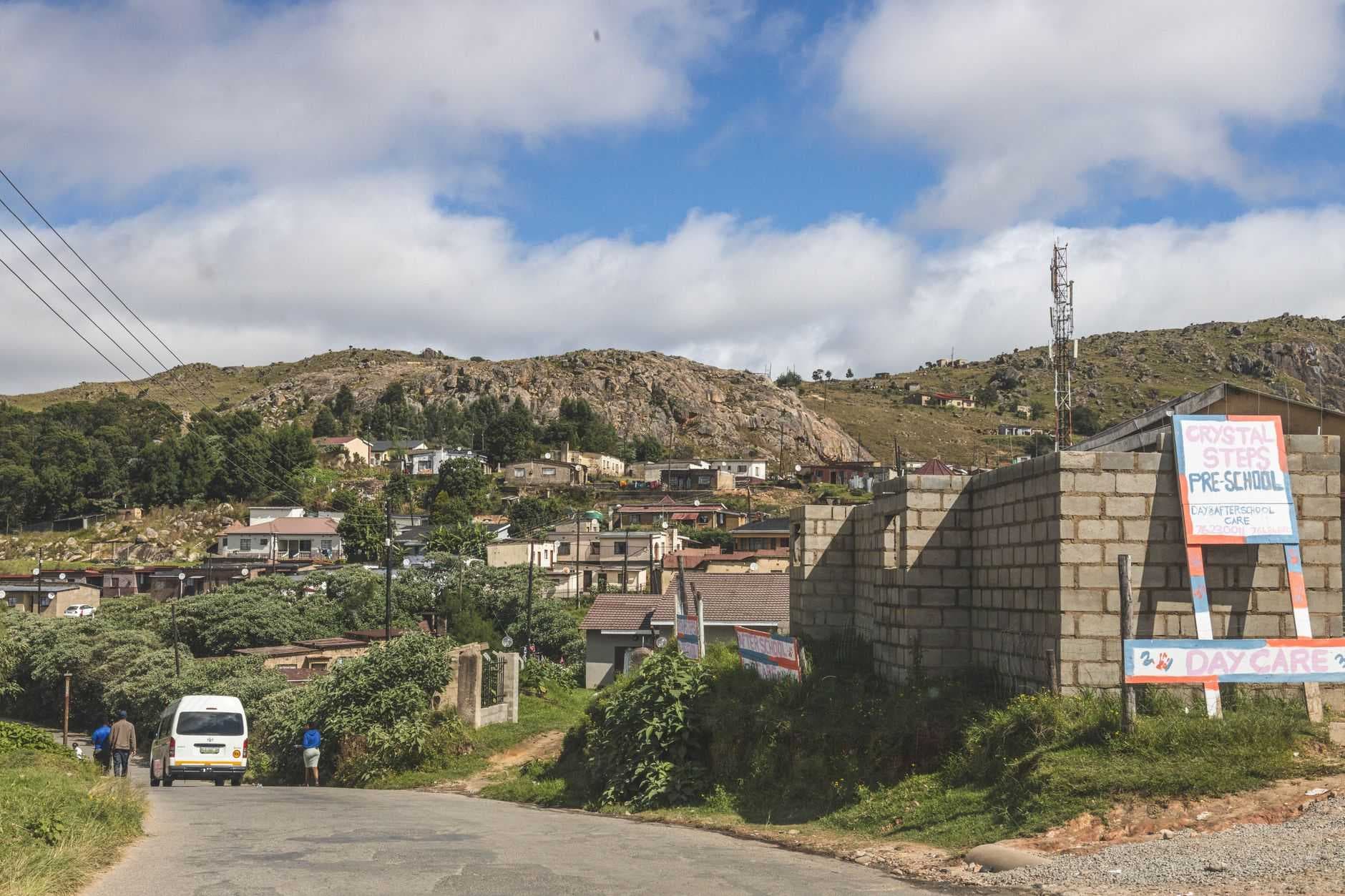 A paved road winds through a rugged Highveld landscape near Piggs Peak, featuring scattered hillside homes, rocky terrain, and a roadside preschool sign.