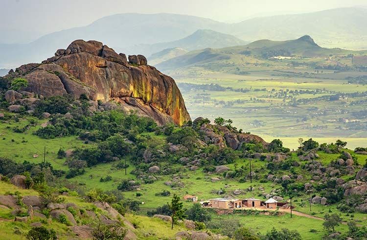 Rural countryside landscape with a large rocky outcrop, green hills, scattered boulders, and distant mountains under a hazy sky in the Swaziland region near Sid