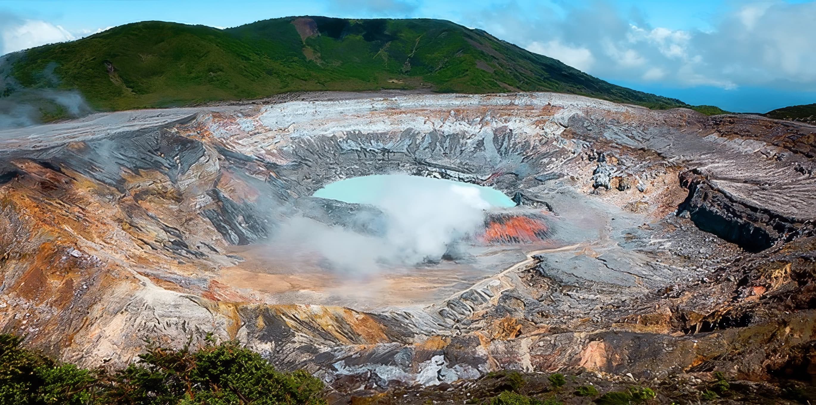 Volcanic crater with turquoise lake, steam vents, mineral-stained slopes, and green highland ridges under a cloudy sky.