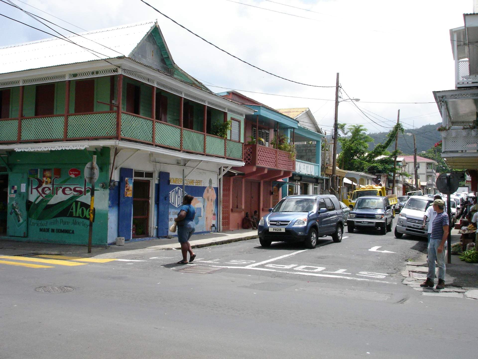 Street scene in a Caribbean town near Mahaut with colorful two-story buildings, parked vehicles, pedestrians, and a 'Made in Dominica' mural.