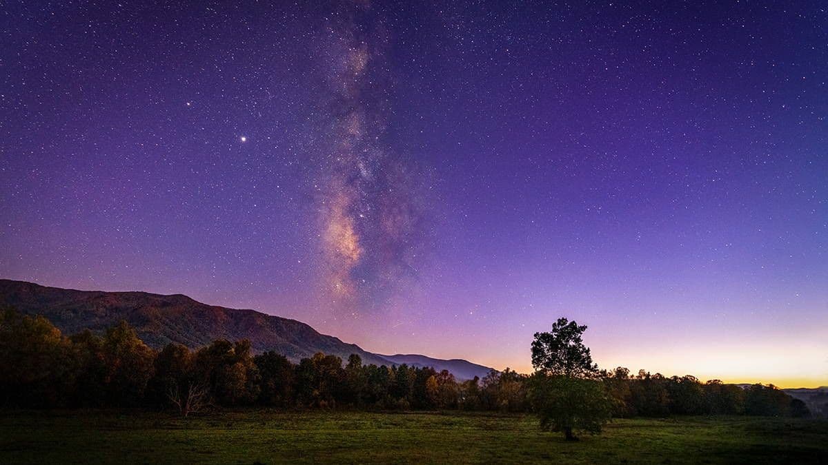 Night sky with stars and the Milky Way over a rural landscape with hills, trees, and a grassy field near Dubreuil, Mauritius.