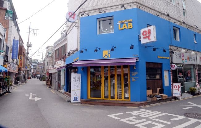 A bright blue corner shop with yellow-framed windows and 'ROASTING LAB' signage on a narrow commercial street with Korean storefronts.