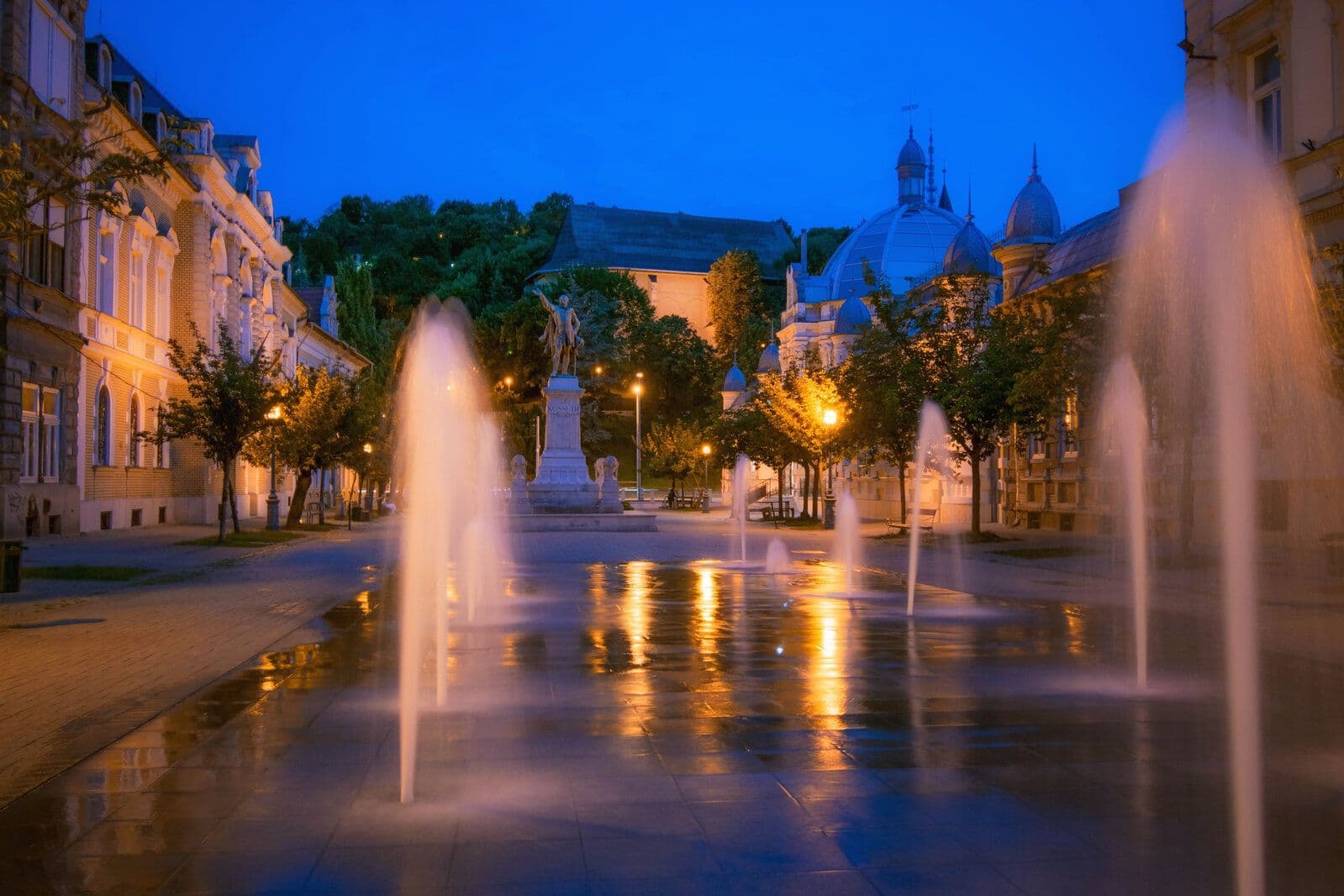 Night view of Petofi Square in Debrecen featuring illuminated fountains, a central statue, and historic architecture under a blue sky.