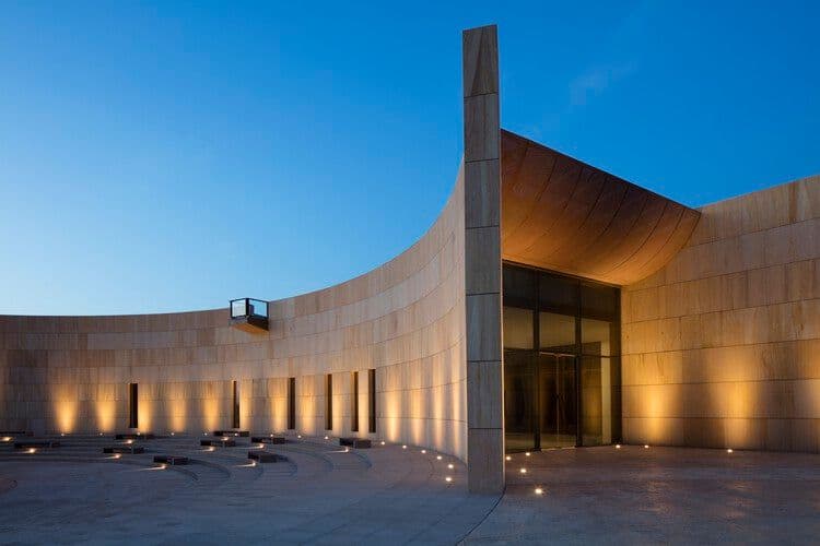 Modern curved stone architecture of the Mleiha Archaeological Centre illuminated at twilight, featuring a sweeping wall and glass entrance.