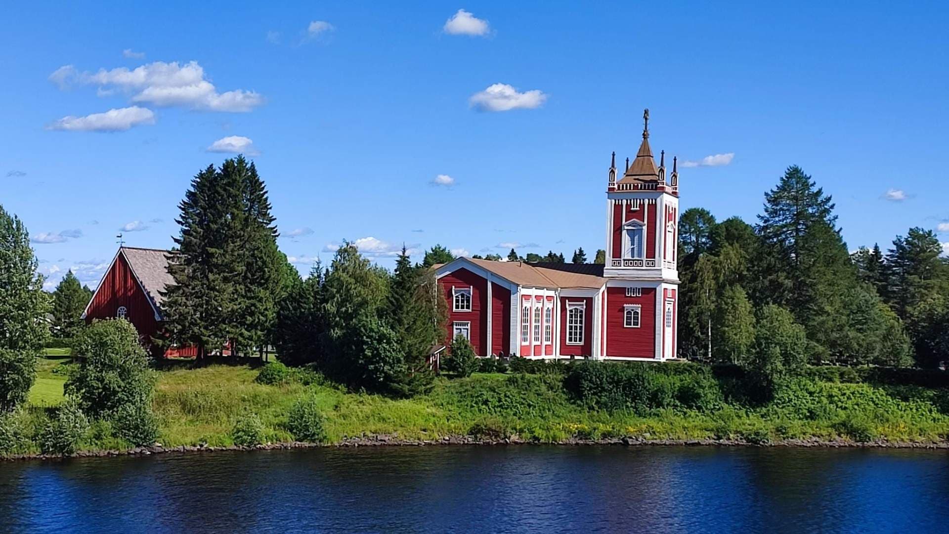 A red wooden church with a white tower stands on a grassy bank beside a river under a blue sky with scattered clouds.