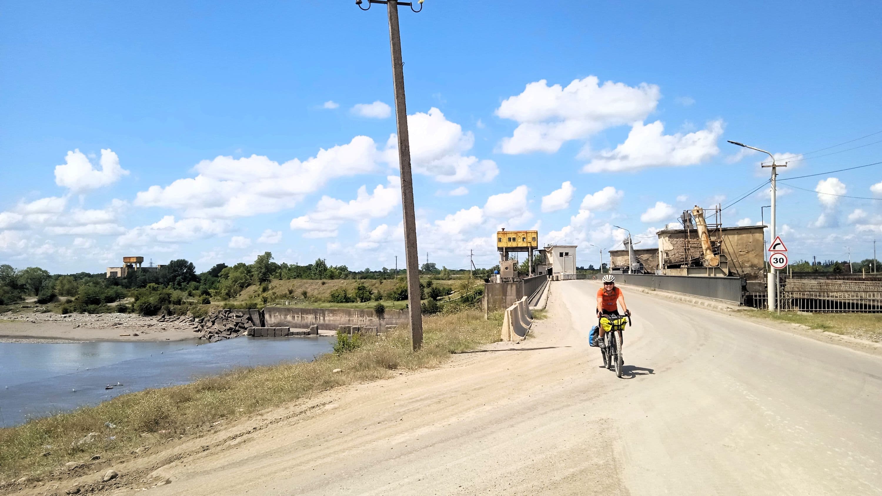 A cyclist rides a gravel road near a river and industrial structures under a blue sky, illustrating transport infrastructure.