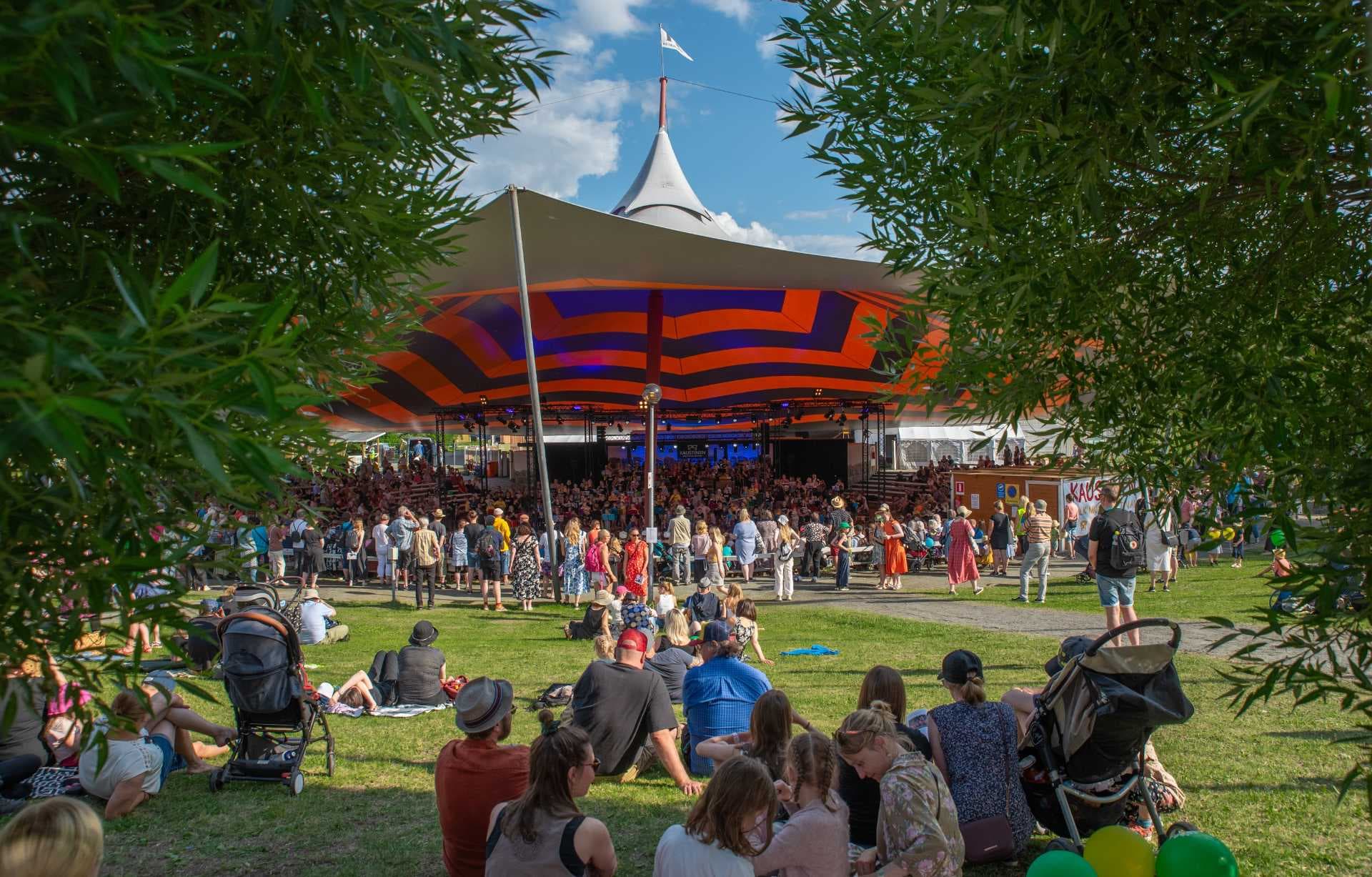 Crowds gather on a grassy lawn before a large, colorful striped festival tent stage under a blue sky, framed by green tree branches at the Kaustinen Folk Music