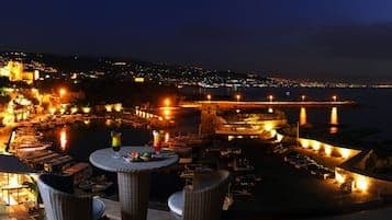 Nighttime rooftop lounge with tables overlooking a coastal village and harbor, illuminated by warm lights.