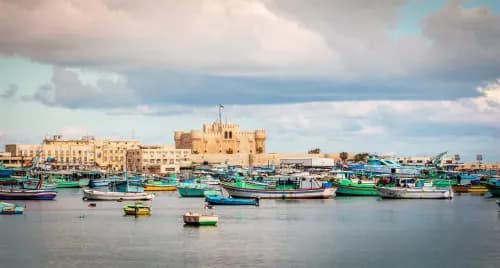 Coastal harbor with colorful fishing boats and a historic stone fortress on the waterfront under a cloudy sky.