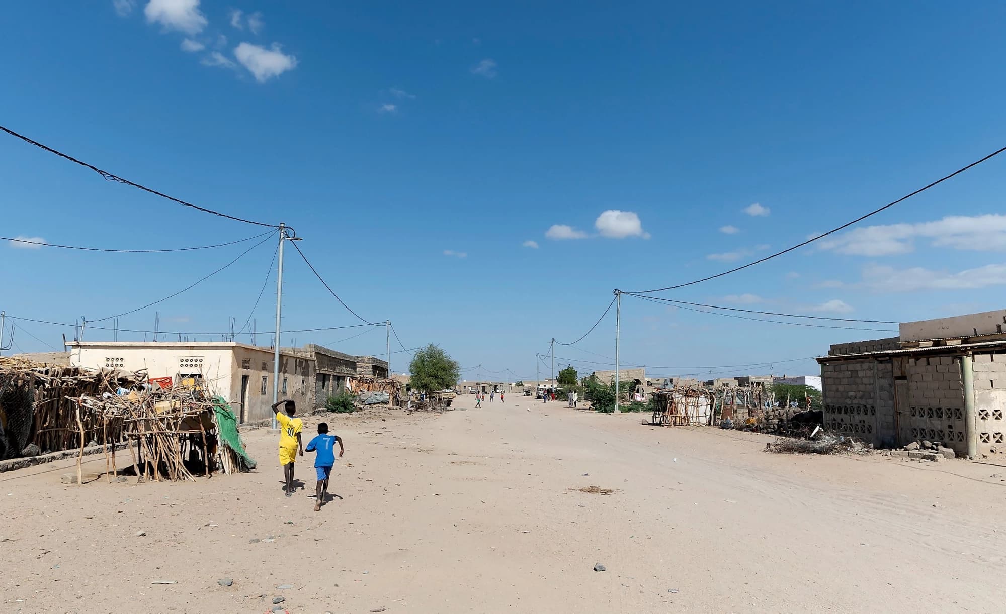 Wide unpaved road in a dusty settlement under a blue sky, with two children running away from the camera and simple block structures lining the street.