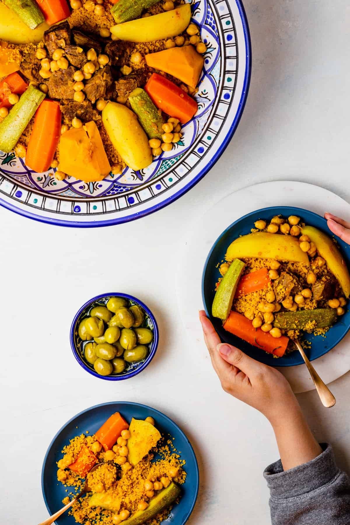 Overhead shot of couscous with vegetables and chickpeas on patterned plates, with hands serving the dish