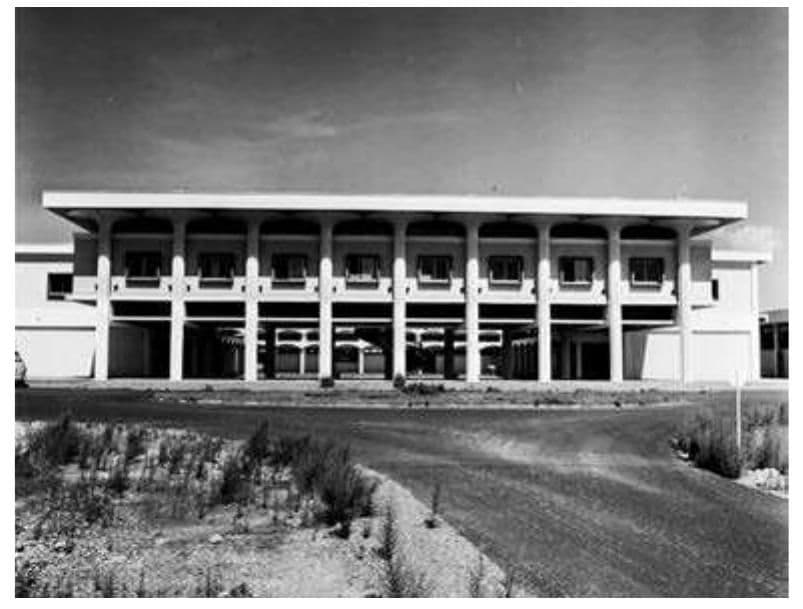 Black-and-white photograph of a two-story building with a colonnaded facade, set against a clear sky with a foreground road and sparse vegetation in Labé, Guine