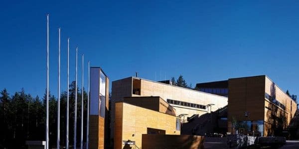 Modern wooden building exterior with tall flagpoles against a clear blue sky, likely the Folk Arts Centre in Kaustinen.