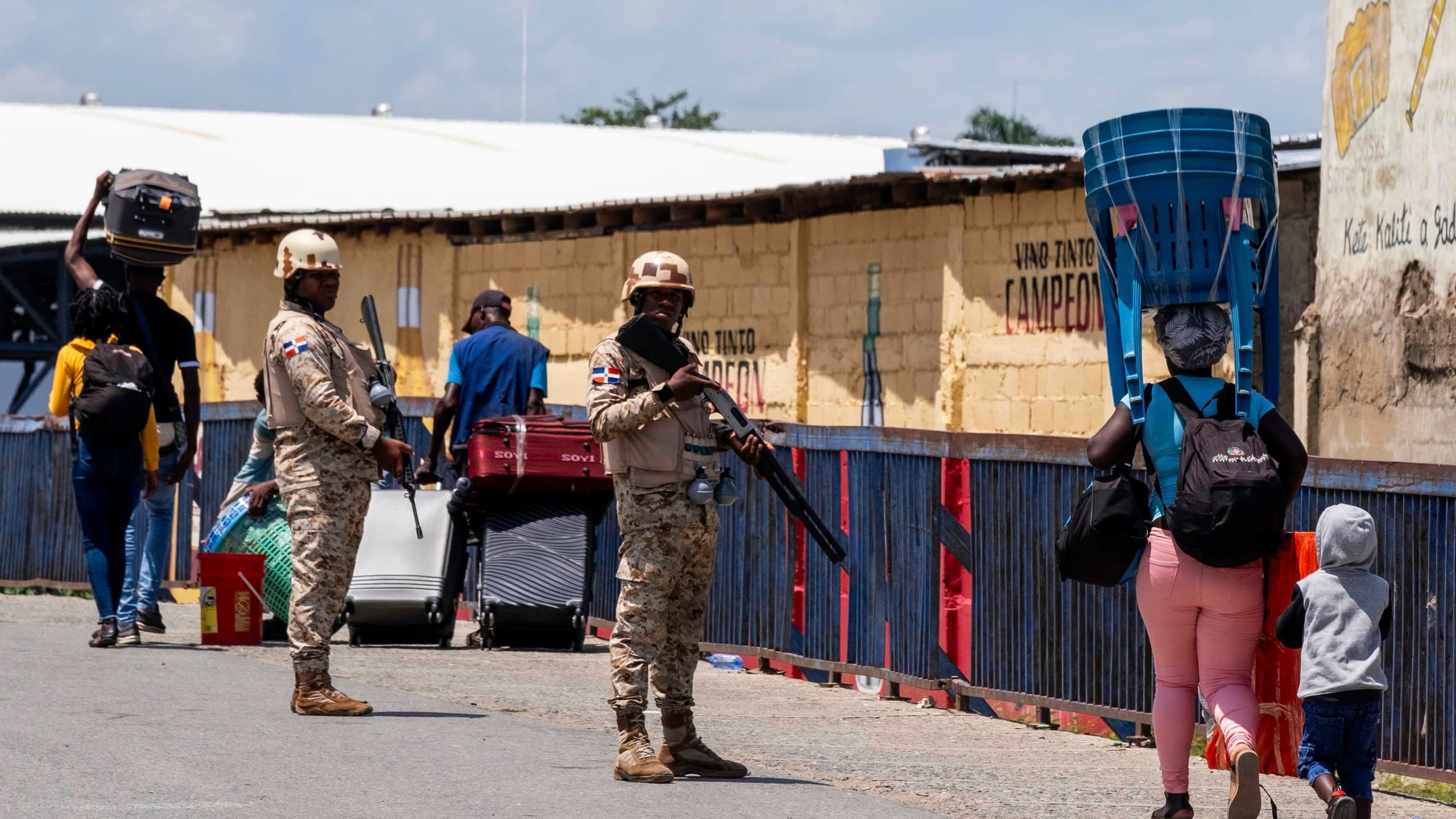 Armed soldiers stand guard on a street in Dajabon while civilians carry luggage and goods past a yellow building.