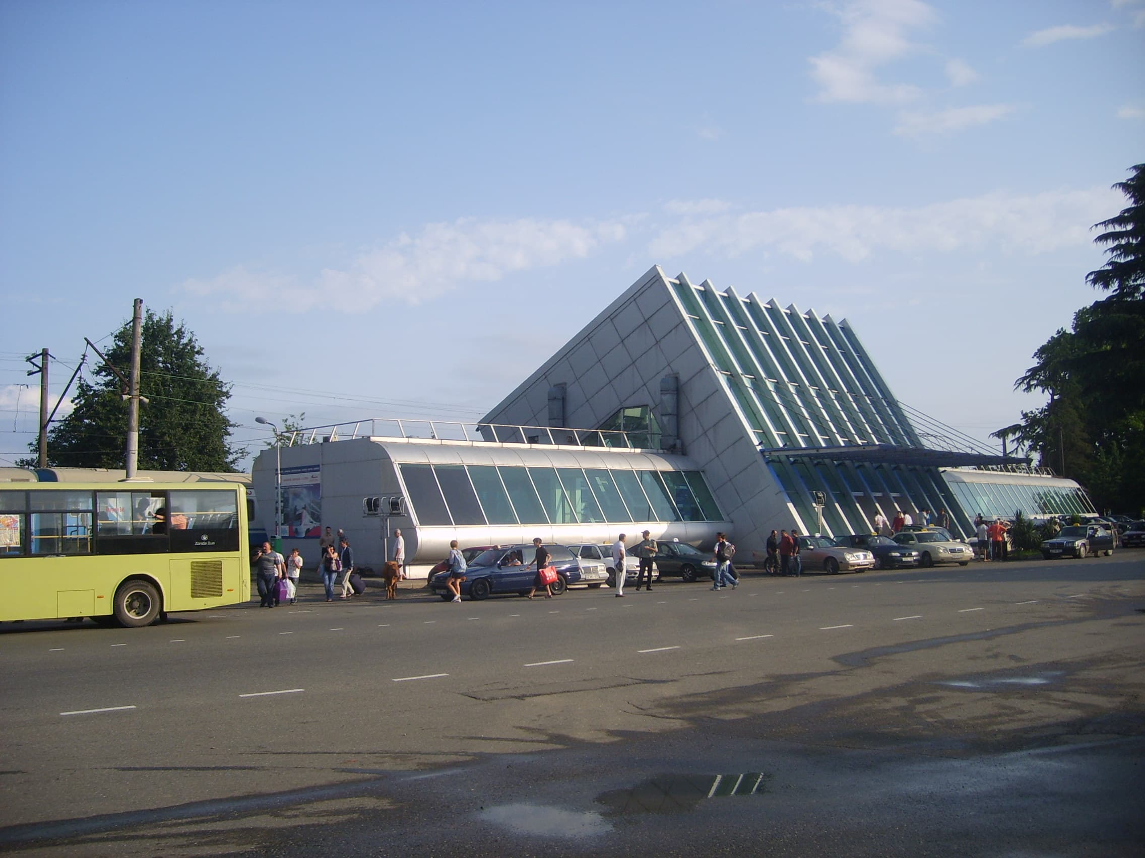 Exterior view of the Makhinjauri Railway Station featuring a distinctive angular glass and concrete structure, with a yellow bus and passengers on the street.
