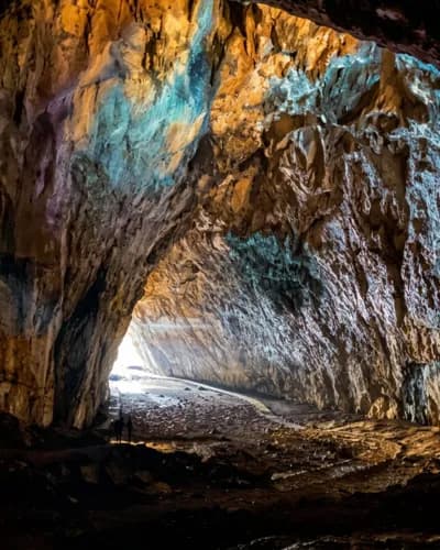 A view from inside a rugged cave looking out toward a bright opening, featuring textured rock walls illuminated by warm and cool artificial lighting.