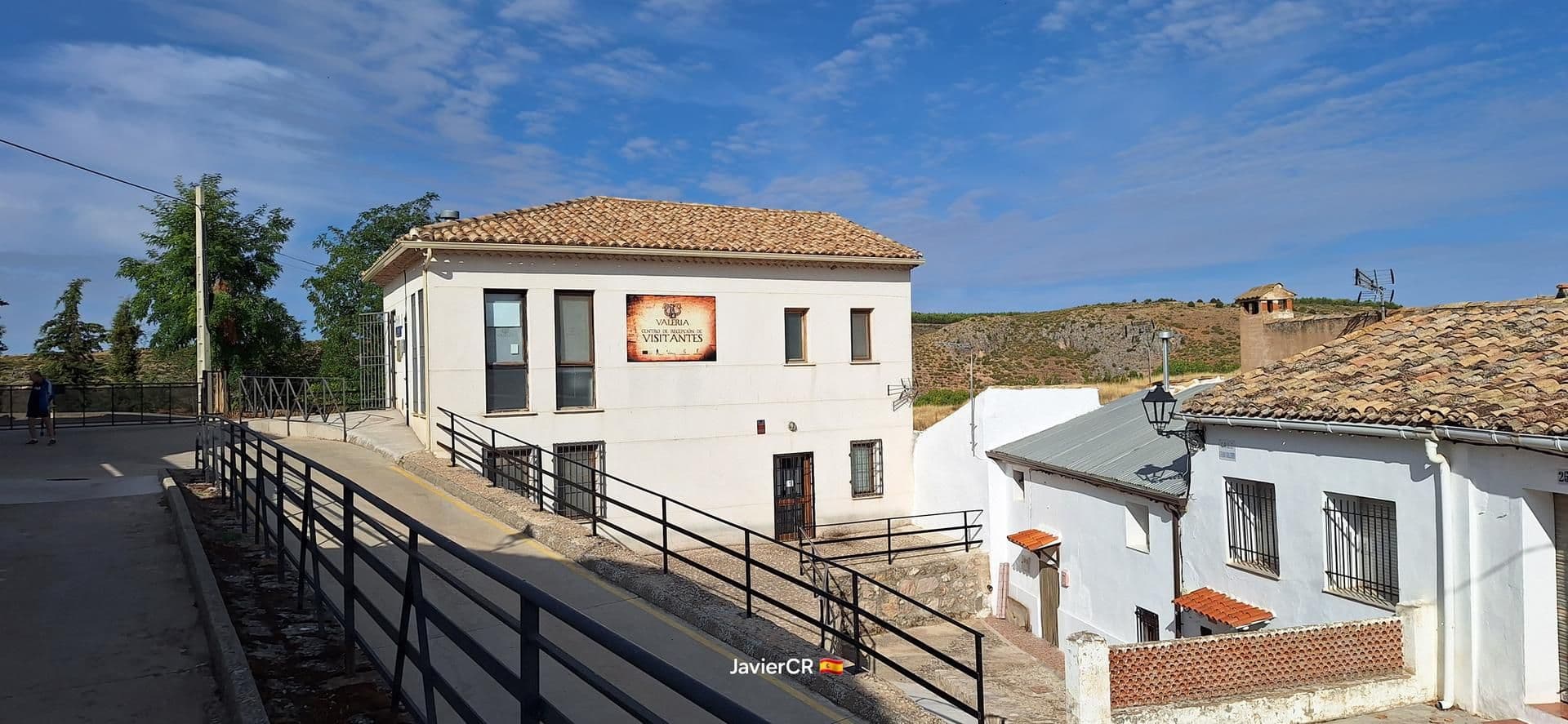 Wide view of a small rural village with white buildings, tiled roofs, and a visitor center sign, set against a hilly Castilian landscape under a blue sky.