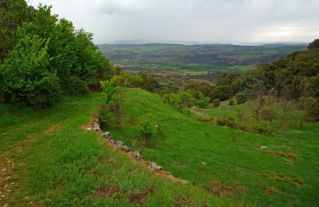 Green rural hillside with a grassy path and stone border, overlooking a wide valley landscape under an overcast sky.
