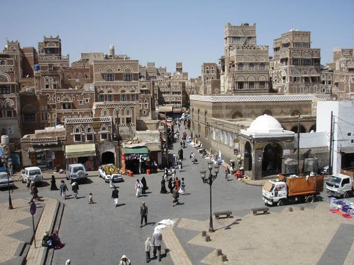 Wide-angle view of a bustling town square in As Sawm, featuring traditional mud-brick architecture, pedestrians, and vehicles under a clear blue sky.