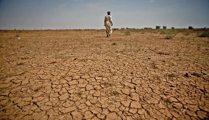 A lone figure walks across a vast, cracked, arid landscape under a clear sky, illustrating the harsh heat and dry terrain of the Sahel.