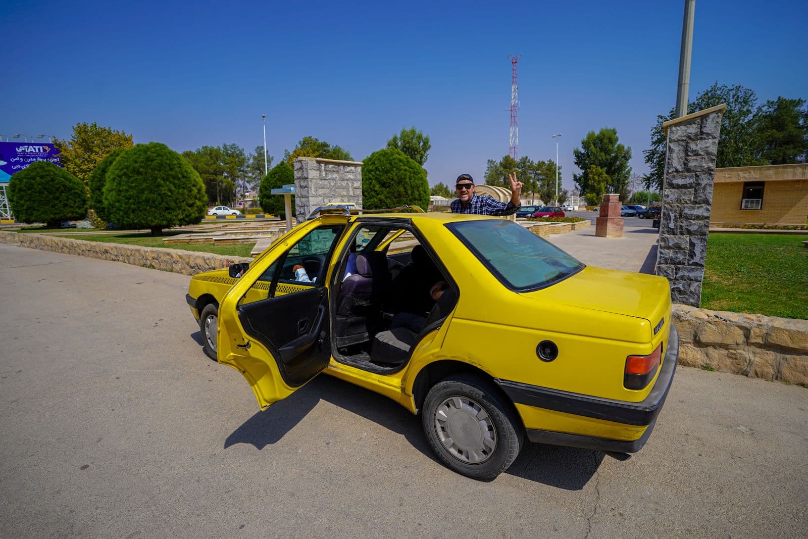 Yellow taxi on a paved roadside in Iraq with trees and stone pillars in the background, illustrating local road transport.