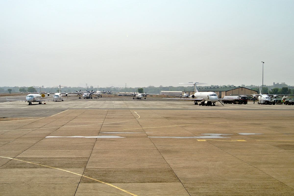 Wide view of a small airport apron with several parked aircraft, ground vehicles, and hangars under a hazy sky.